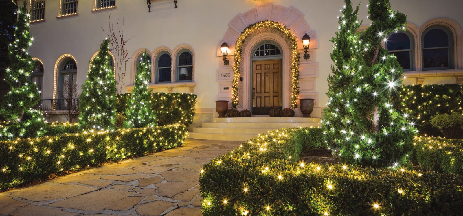 Warm white holiday lights on an entry walk, hedges, and small evergreens